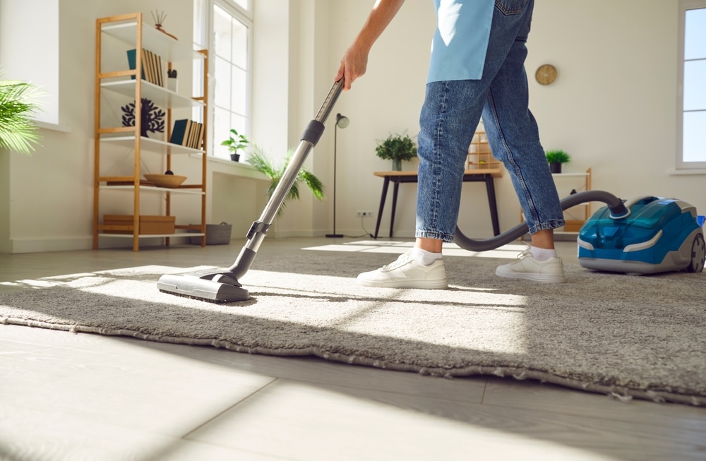 Person vacuuming a gray area rug in a bright living room with wooden bookshelves