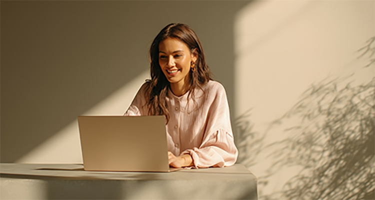 A young woman smiles while working on a laptop at a desk in a sunlit room.