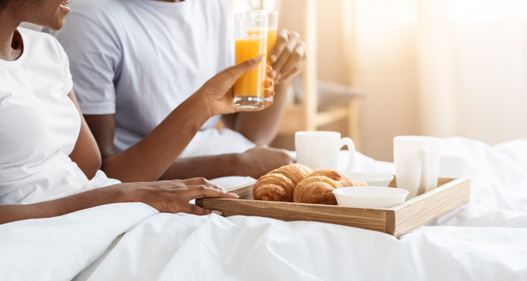 A couple sitting in white bedding holding glasses of orange juice with a tray of croissants and coffee in front of them.