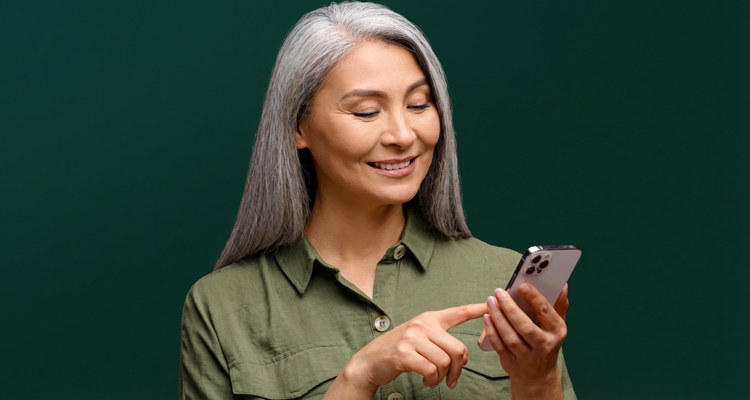 An older woman with grey hair smiles while looking at her smartphone against a green background.