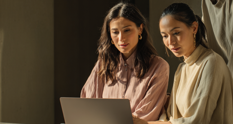 Two professional women, one with long brown hair and one with dark hair pulled back, leaning over a laptop together in soft, warm lighting.