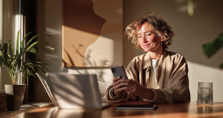 A smiling woman with curly hair looking at her smartphone while sitting at a desk with a laptop, bathed in warm sunlight.